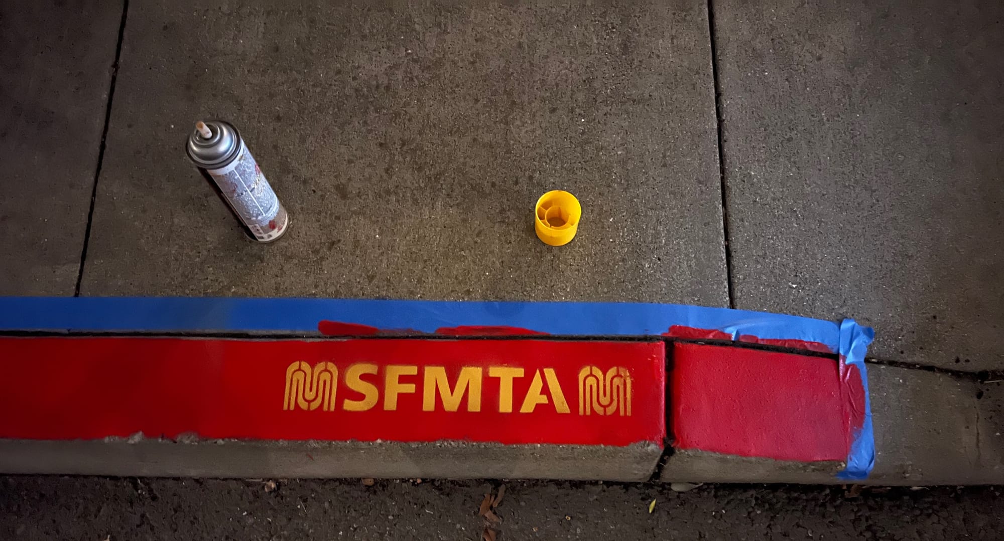 Freshly painted red curb lined with blue painters tape and a yellow SFMTA logo spray-painted on it